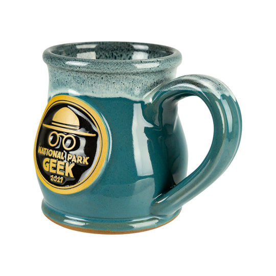 A teal ceramic mug with a speckled glaze and a large handle, featuring a circular gold and black National Park Geek 2027 logo on the front, set against a plain white background.