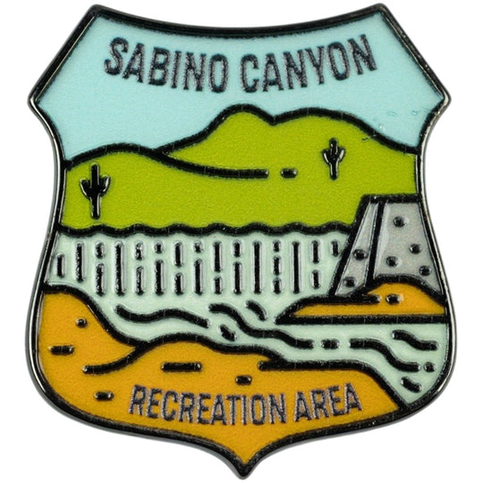 A shield-shaped enamel pin featuring a stylized landscape labeled &