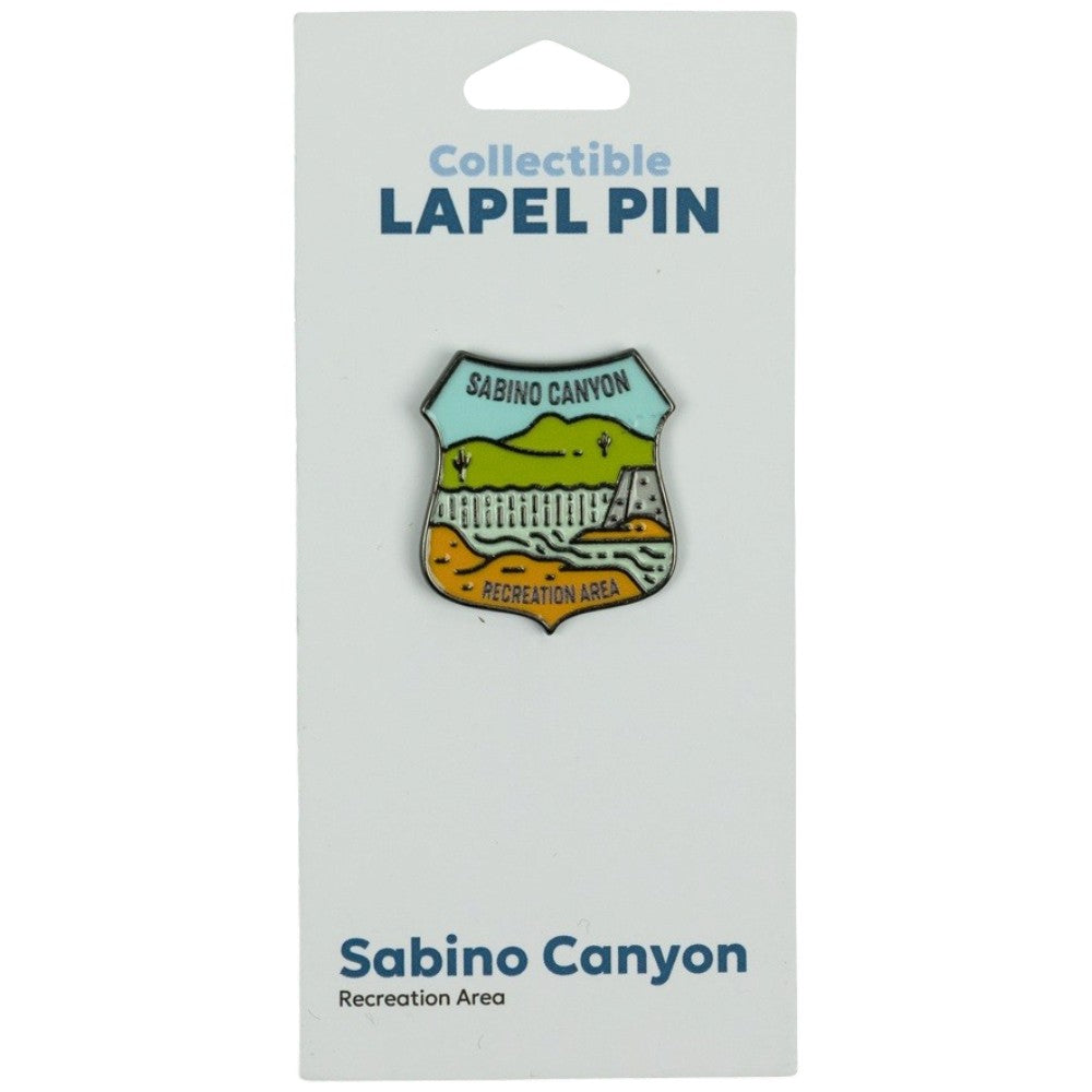 A Sabino Canyon Recreation Area collectible lapel pin displayed on its white backing card. The shield-shaped pin features a multicolor landscape design with green hills, a white fence, and orange/yellow ground, with &