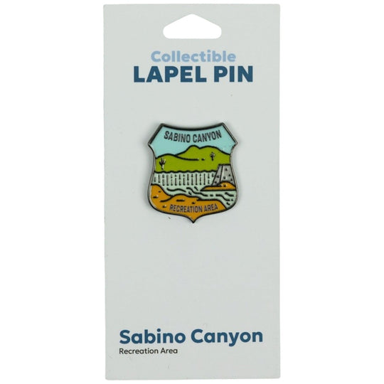 A Sabino Canyon Recreation Area collectible lapel pin displayed on its white backing card. The shield-shaped pin features a multicolor landscape design with green hills, a white fence, and orange/yellow ground, with &