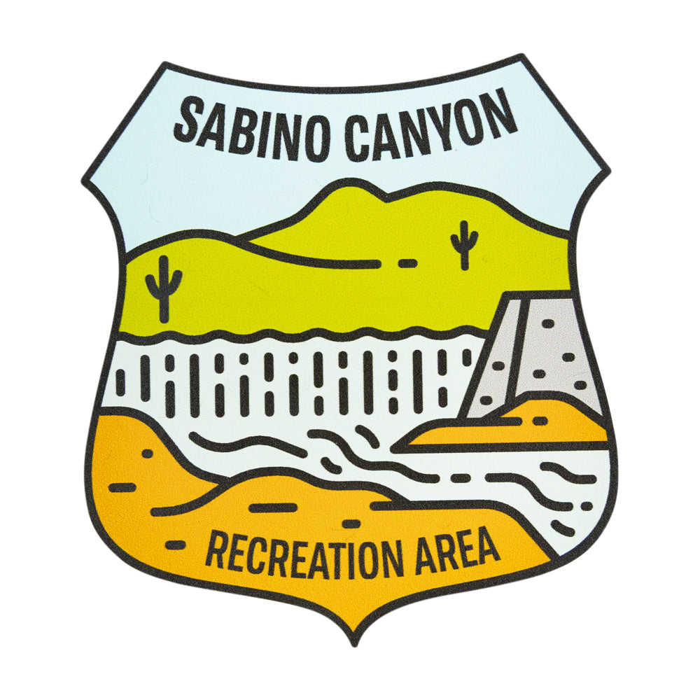 A shield-shaped illustrated sticker reading “SABINO CANYON” at the top and “RECREATION AREA” at the bottom, showing stylized desert hills, cacti, a dam, and flowing lines in yellow, orange, white, and light blue on a black outline.