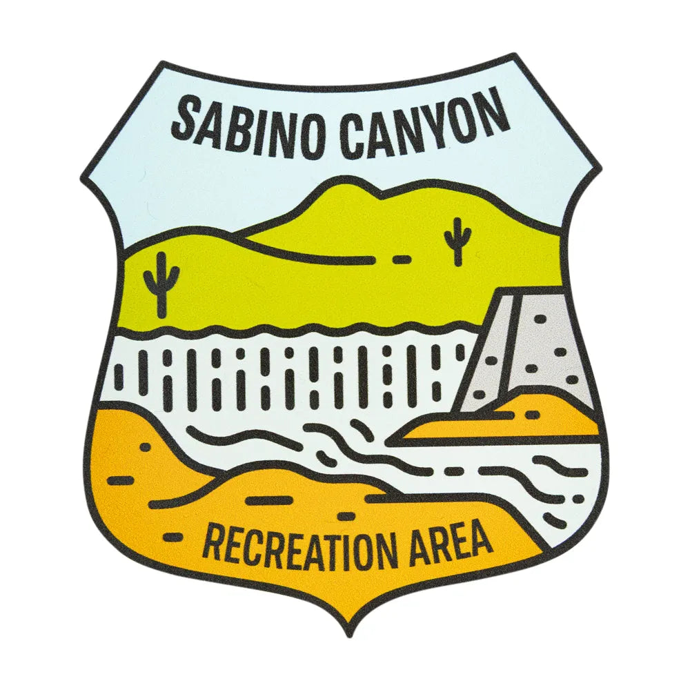 A shield-shaped illustrated sticker reading “SABINO CANYON” at the top and “RECREATION AREA” at the bottom, showing stylized desert hills, cacti, a dam, and flowing lines in yellow, orange, white, and light blue on a black outline.