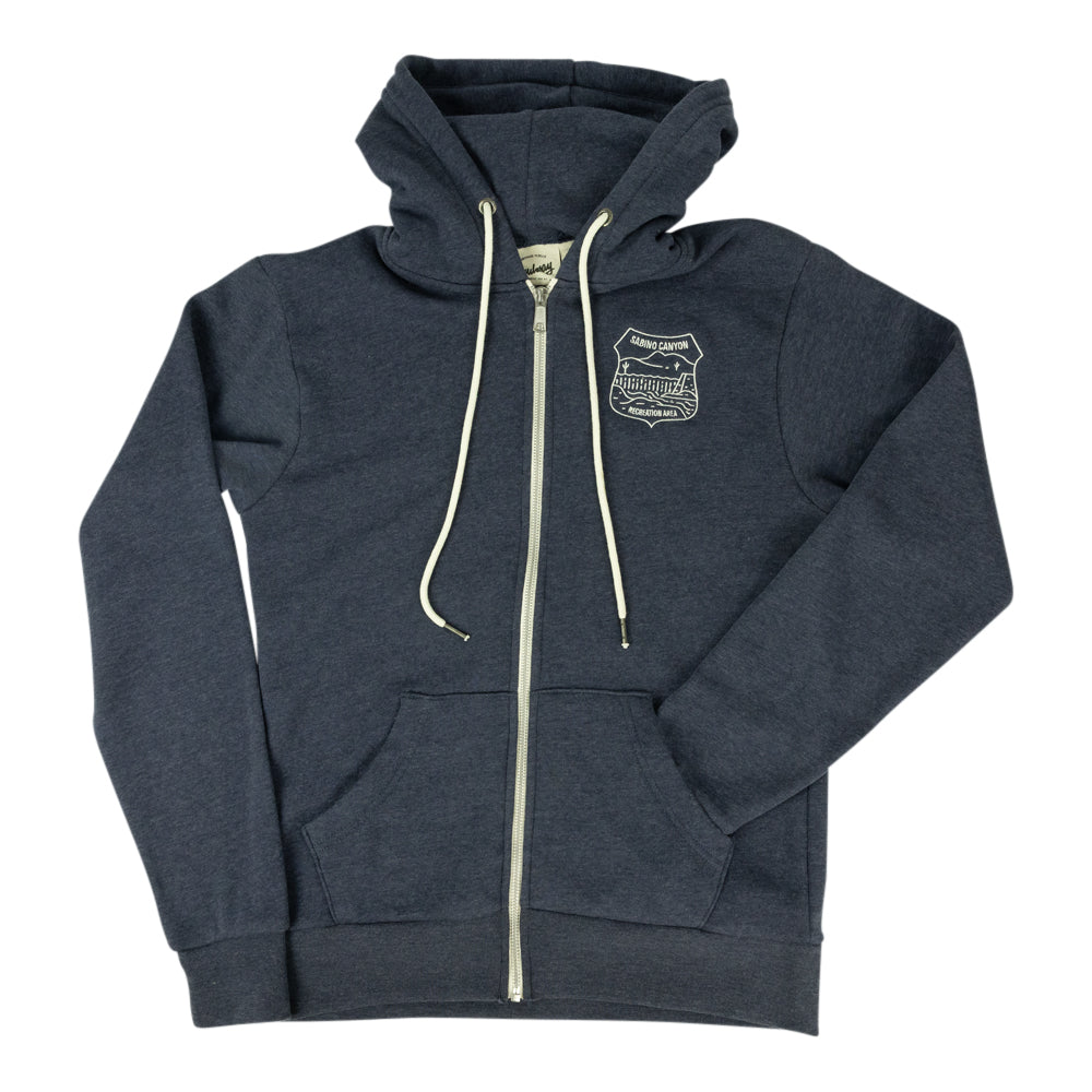 A navy blue zip-up hoodie with cream-colored drawstrings and a zipper, featuring a small embroidered shield logo on the left chest. The hoodie is laid flat against a solid white background.