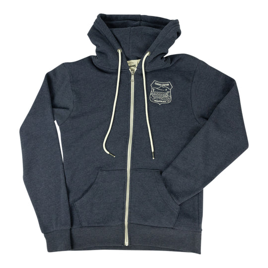 A navy blue zip-up hoodie with cream-colored drawstrings and a zipper, featuring a small embroidered shield logo on the left chest. The hoodie is laid flat against a solid white background.