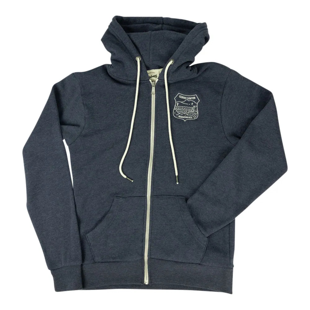 A navy blue zip-up hoodie with cream-colored drawstrings and a zipper, featuring a small embroidered shield logo on the left chest. The hoodie is laid flat against a solid white background.