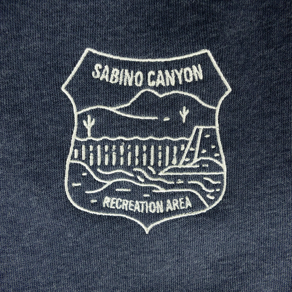 Close-up of a dark navy blue fabric with a white embroidered shield-shaped graphic reading &