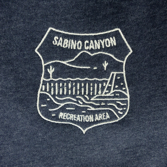 Close-up of a dark navy blue fabric with a white embroidered shield-shaped graphic reading &