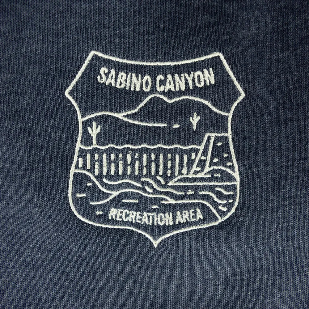 Close-up of a dark navy blue fabric with a white embroidered shield-shaped graphic reading &