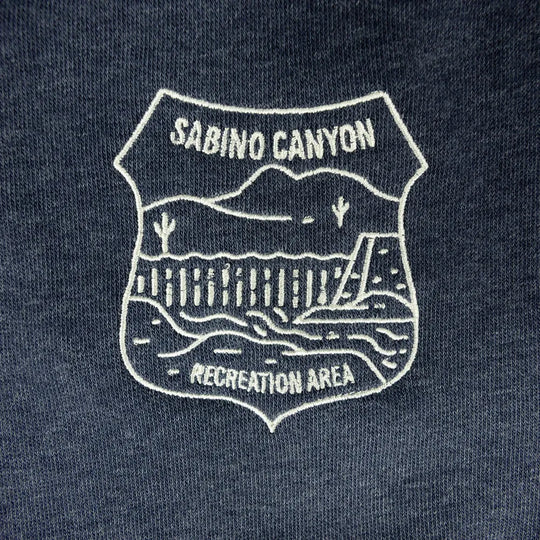 Close-up of a dark navy blue fabric with a white embroidered shield-shaped graphic reading &