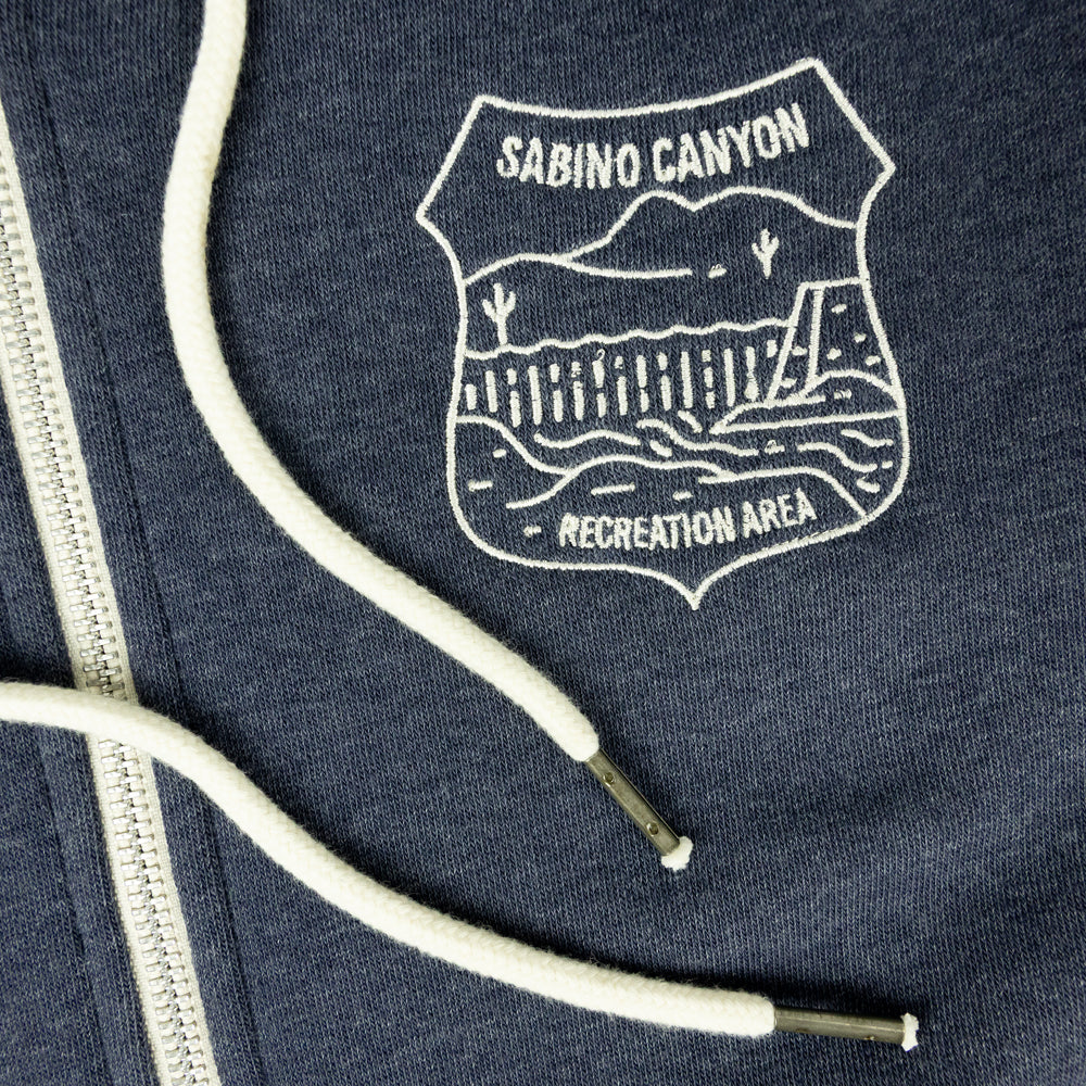 Close-up of a navy blue zippered hoodie featuring a white embroidered shield logo that reads &