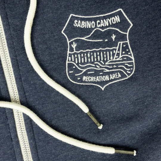 Close-up of a navy blue zippered hoodie featuring a white embroidered shield logo that reads &