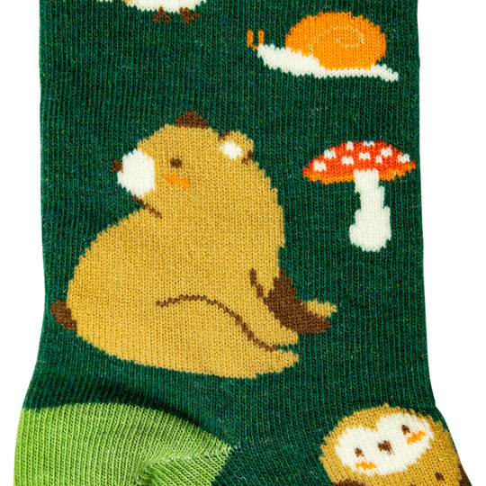 A close-up of a dark green knit sock featuring a playful woodland pattern with a large tan bear, a red-and-white mushroom, a white snail, and a green heel area.