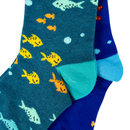 A close-up of a pair of patterned crew socks on a white background. One sock is dark teal with yellow, orange, and light blue fish motifs, and the other is royal blue with a light blue toe and a small orange fish pattern.