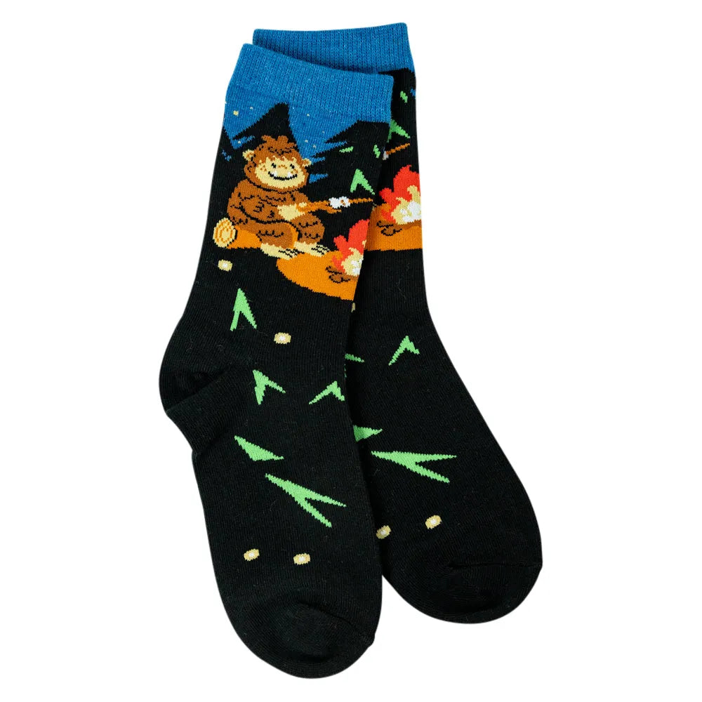 A pair of black crew socks with a colorful Bigfoot camping graphic on the upper leg and green pine tree patterns on the feet, shown against a plain white background.