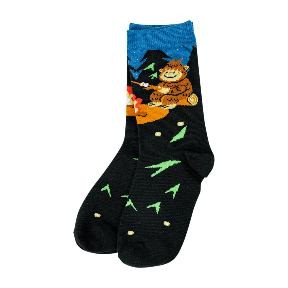 A pair of black crew socks featuring a colorful graphic of a friendly brown bear sitting by a campfire under a blue night sky, with green pine tree silhouettes and small gold stars on a white background.