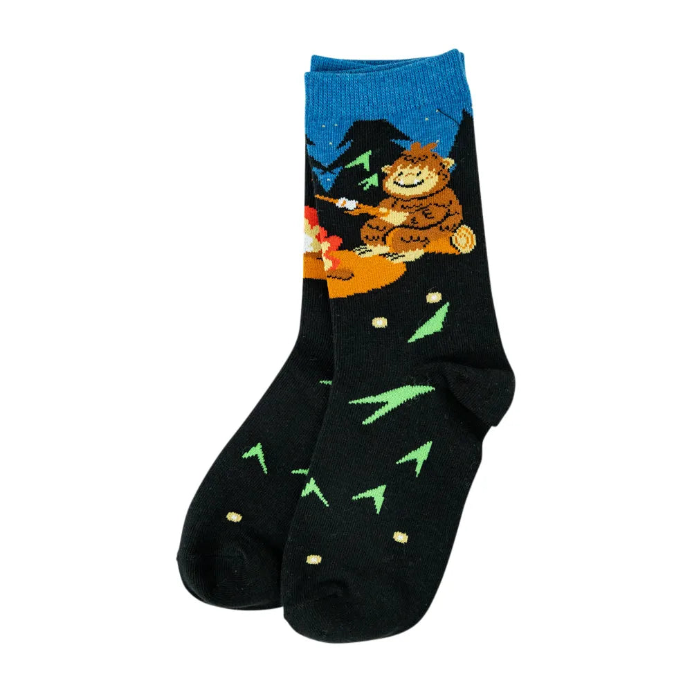 A pair of black crew socks featuring a colorful graphic of a friendly brown bear sitting by a campfire under a blue night sky, with green pine tree silhouettes and small gold stars on a white background.