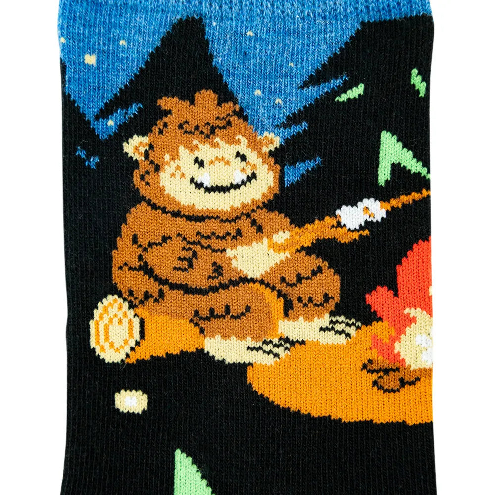 A close-up of a knitted sock featuring a cartoon bigfoot roasting marshmallows over a campfire at night under a blue starry sky.