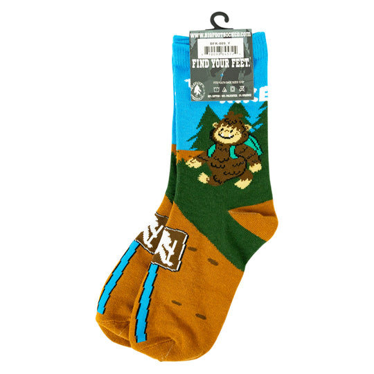 A pair of novelty crew socks featuring a cartoon Bigfoot design on a blue, green, and brown background, with a hangtag attached at the top.