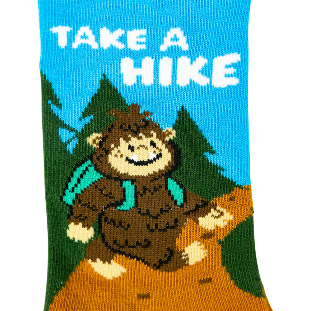 A single knit sock featuring a cartoon brown bear wearing a teal backpack and scarf, standing on an orange path with green pine trees and a bright blue sky. The words "TAKE A HIKE" are knitted across the top in white.
