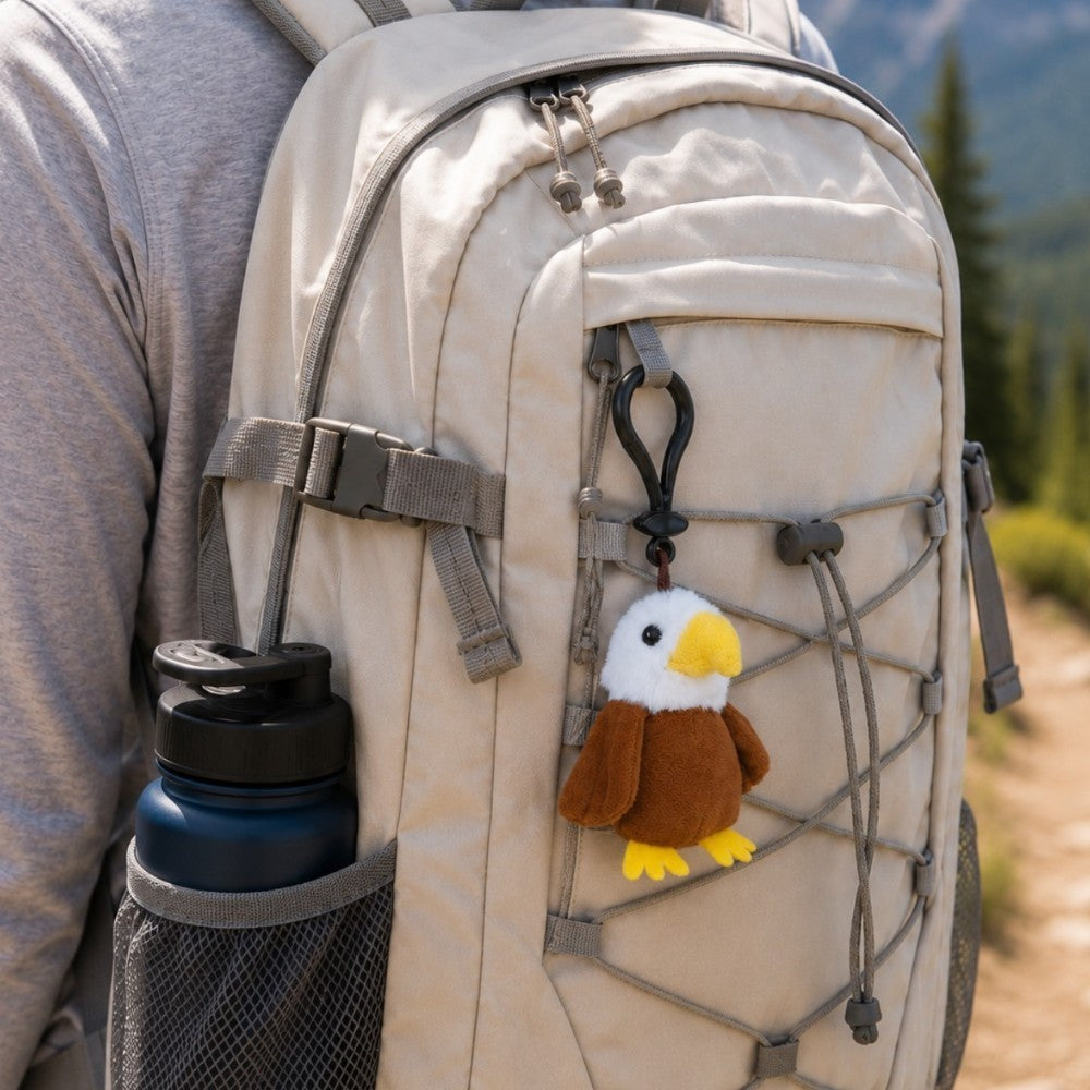 A close-up of a beige hiking backpack worn by a person outdoors, with a small plush eagle keychain hanging from the front webbing. The backpack has gray straps, a side water bottle pocket with a dark blue bottle, and a blurred forest trail in the background.