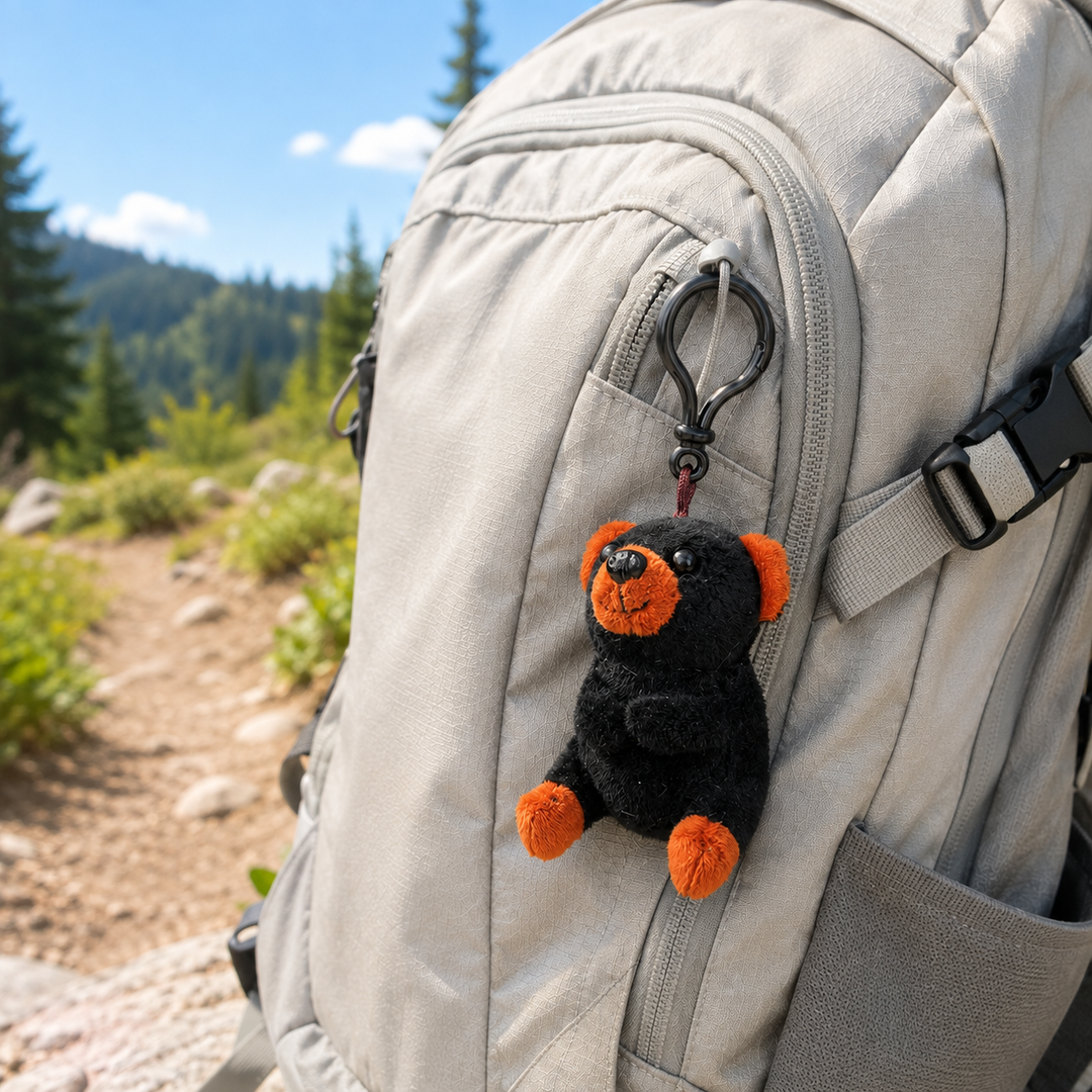 A small plush black bear keychain with orange paws and snout hanging from a black carabiner on the front pocket of a light gray hiking backpack, photographed outdoors on a trail with trees and blue sky in the background.