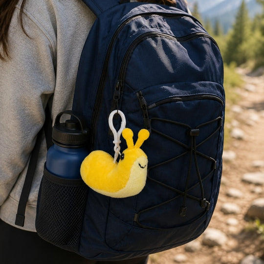 A navy blue hiking backpack worn by a person outdoors, with a yellow plush snail keychain hanging from the front pocket and a blue water bottle in the side mesh pocket.