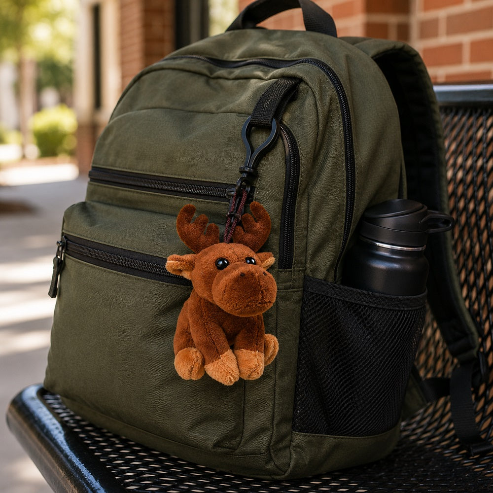 An olive green backpack with black zippers and a side mesh pocket, sitting on a black metal bench. A brown plush moose keychain is hanging from the zipper, and a black water bottle is in the side pocket.