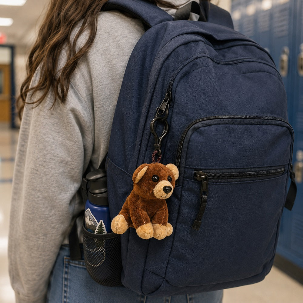 A navy blue backpack worn by a person in a school hallway, featuring a brown plush teddy bear keychain hanging from the zipper.
