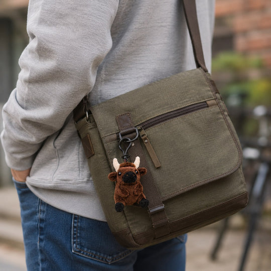 An olive green canvas messenger bag with brown leather trim, worn crossbody by a person in a gray sweatshirt and blue jeans. A small brown plush moose keychain is attached to the zipper.