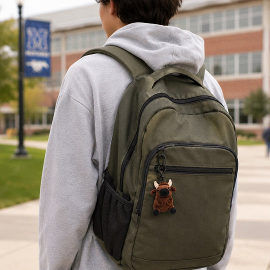 An olive green backpack with black mesh side pockets and a brown bull-shaped keychain charm, worn by a person in a gray hoodie on a college campus walkway.