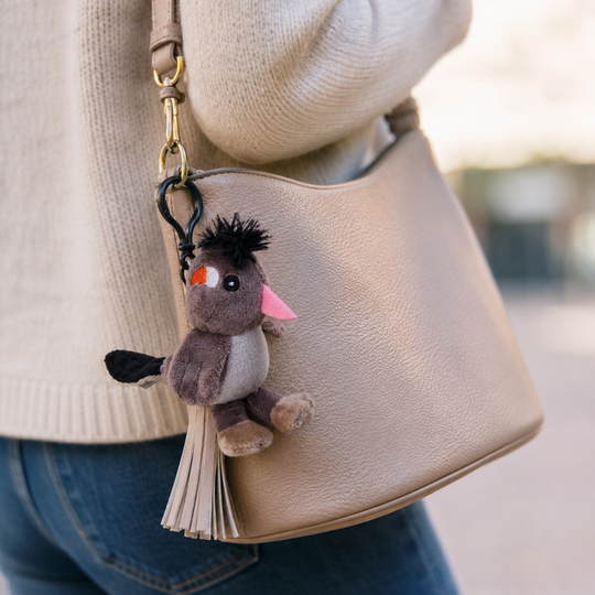 A beige pebbled leather shoulder bag worn by a person in a cream knit sweater and blue jeans, featuring a small plush bird keychain charm with a tassel.