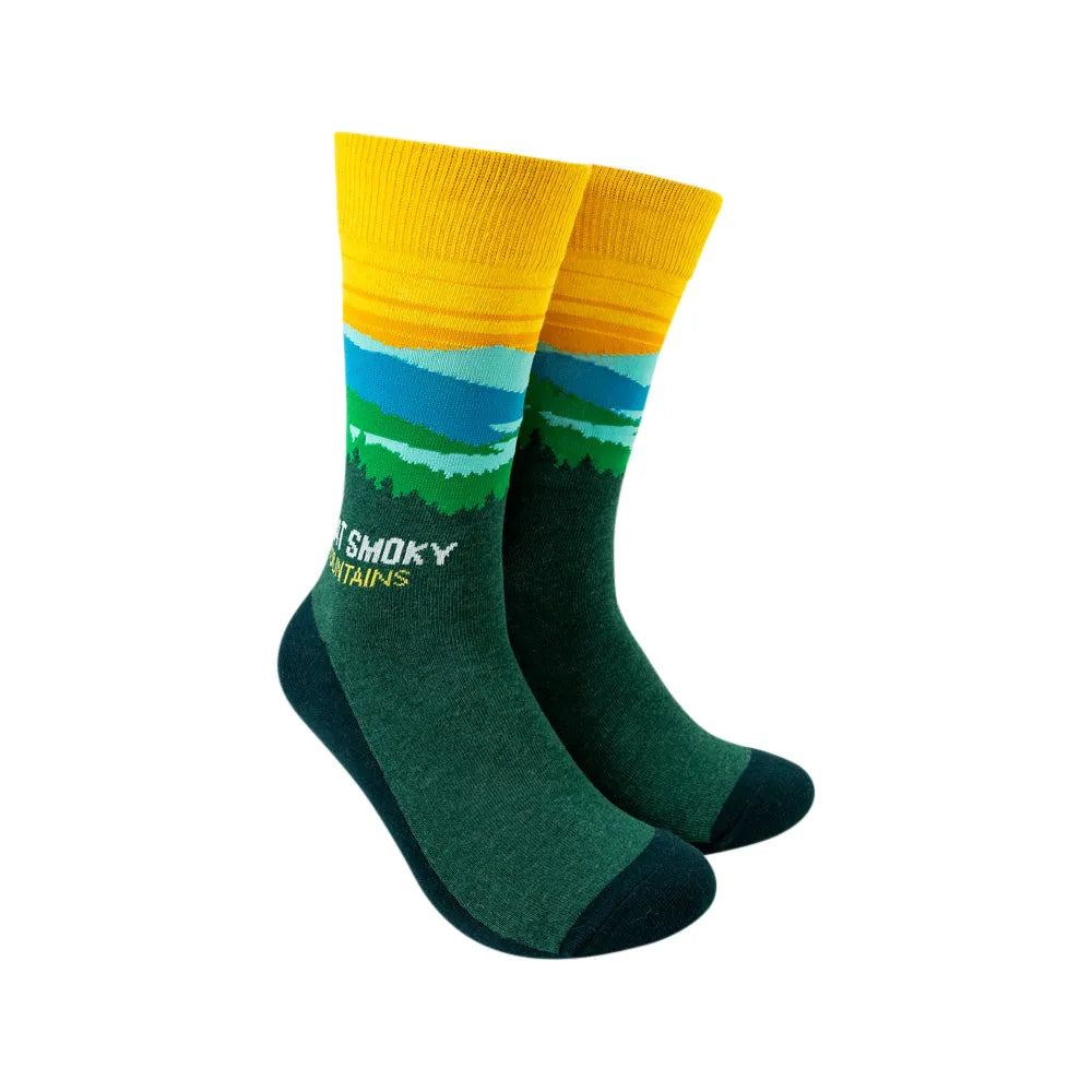 A pair of crew socks with a scenic mountain landscape print, featuring a yellow and orange striped cuff, blue and green hills, and a dark green foot with &
