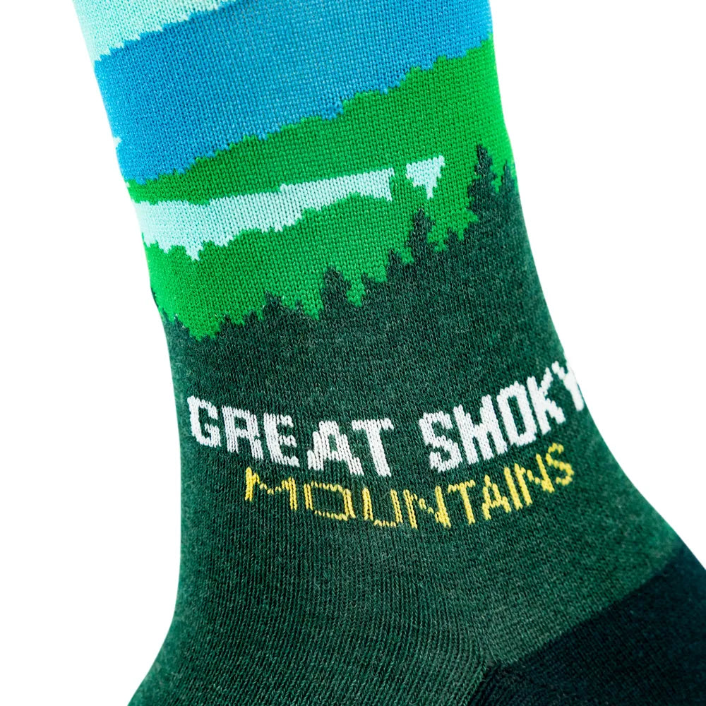 A close-up of a knit sock featuring a stylized mountain landscape with blue sky, green hills, and dark green forest, with the words “GREAT SHOKY MOUNTAINS” printed on the side.