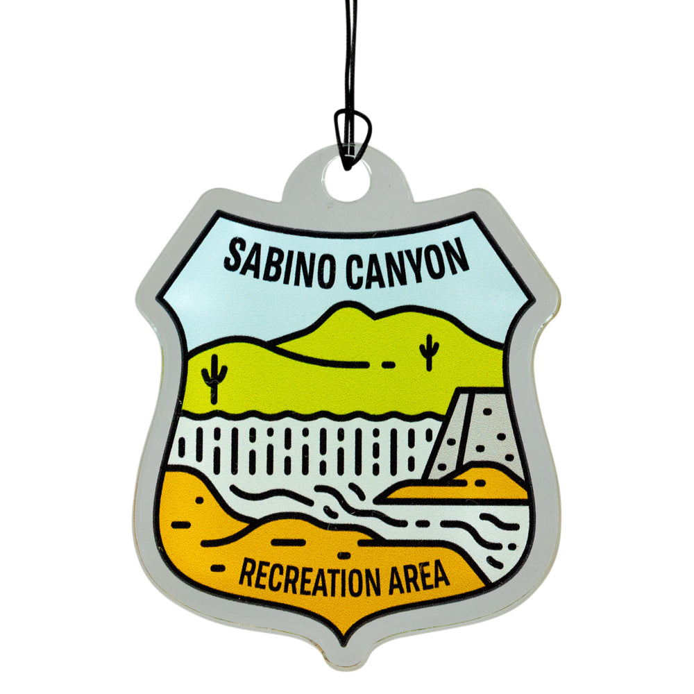 A badge-shaped hanging ornament featuring a stylized desert landscape with the text &