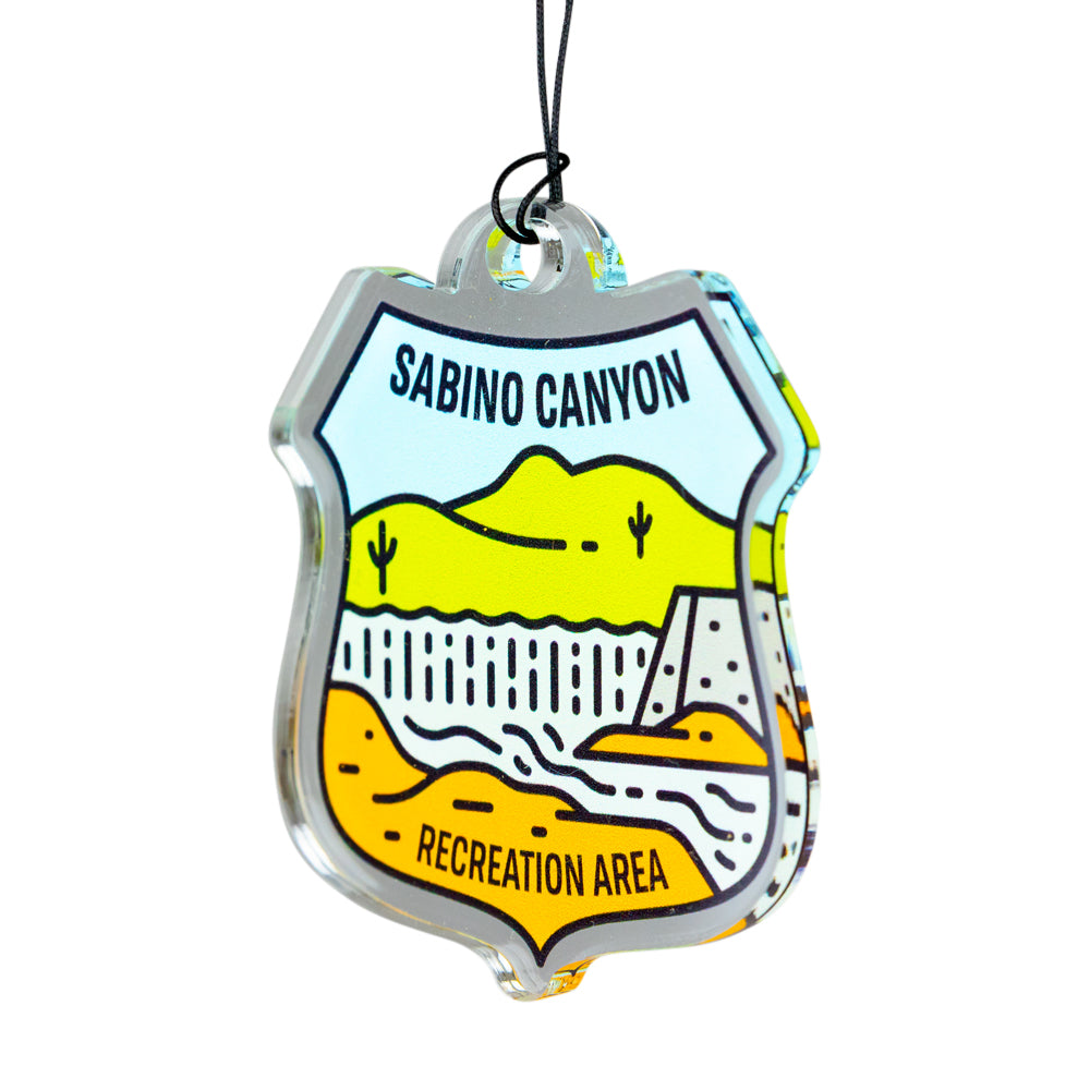 A clear acrylic Sabino Canyon Recreation Area shield-shaped ornament with a black hanging string, featuring a colorful desert landscape illustration.
