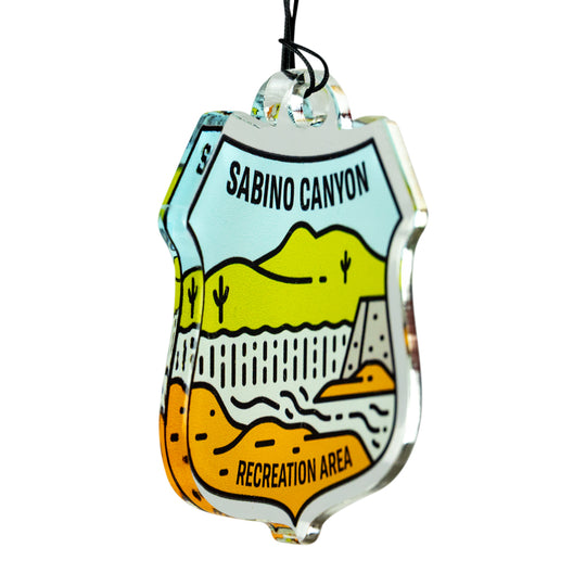 A clear acrylic hanging ornament shaped like a shield, featuring a colorful illustrated desert landscape with the text &
