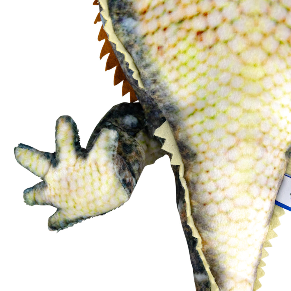 Close-up of a plush gecko toy showing textured yellow-green and cream scales, dark gray feet, and orange-brown spikes against a white background.