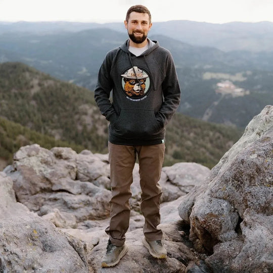 A man standing on a rocky mountain peak wearing a dark charcoal gray hoodie with a graphic of Smokey Bear, paired with brown pants and hiking boots. There is a scenic mountain range in the background.