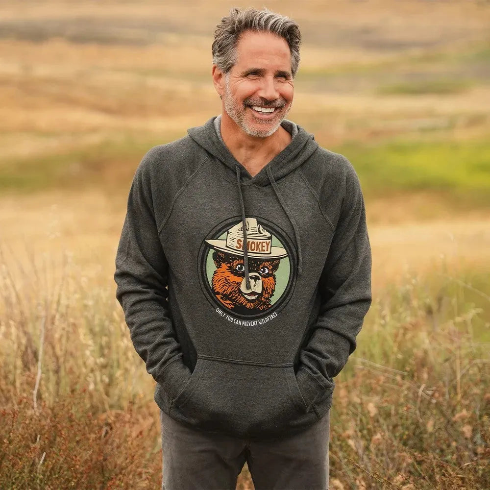 A smiling person with a beard and grey hair standing in prairie grass wearing a dark gray pullover hoodie with a circular graphic of a Smokey Bear on it.