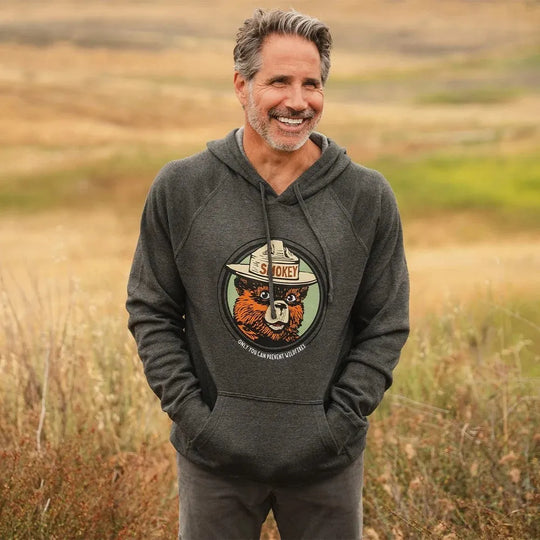 A smiling person with a beard and grey hair standing in prairie grass wearing a dark gray pullover hoodie with a circular graphic of a Smokey Bear on it.