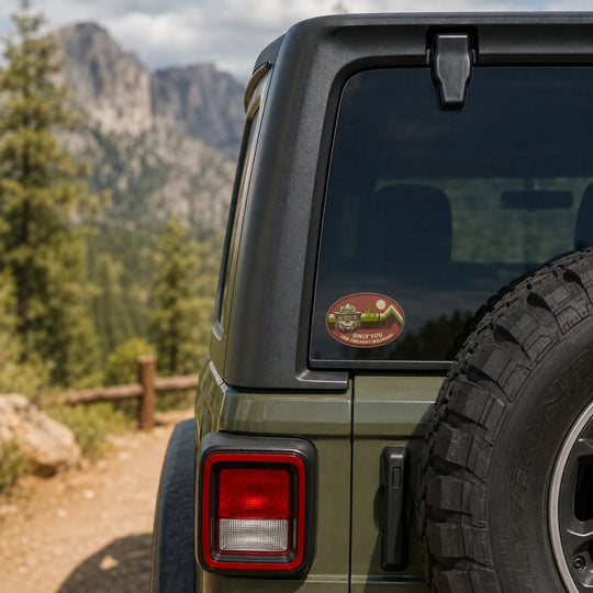 A round red and green Smokey Bear sticker applied to the rear window of an olive green off-road vehicle, with a mountain trail in the background.