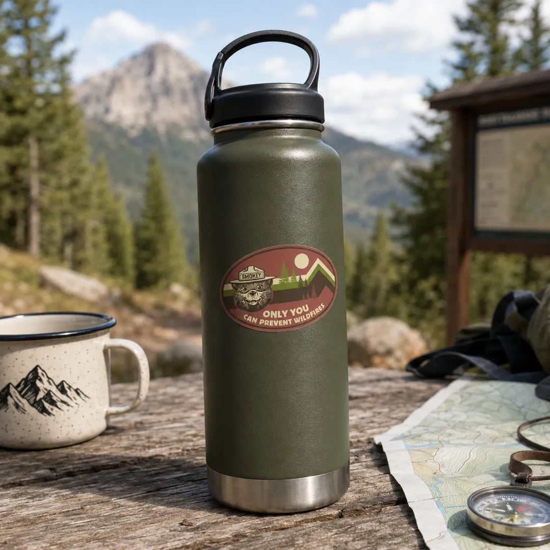 An olive green insulated water bottle with a black lid and handle stands on a wooden table outdoors, with a mountain landscape, pine trees, a mug, and camping items in the background. The bottle has an oval shaped Smokey Bear sticker that says &