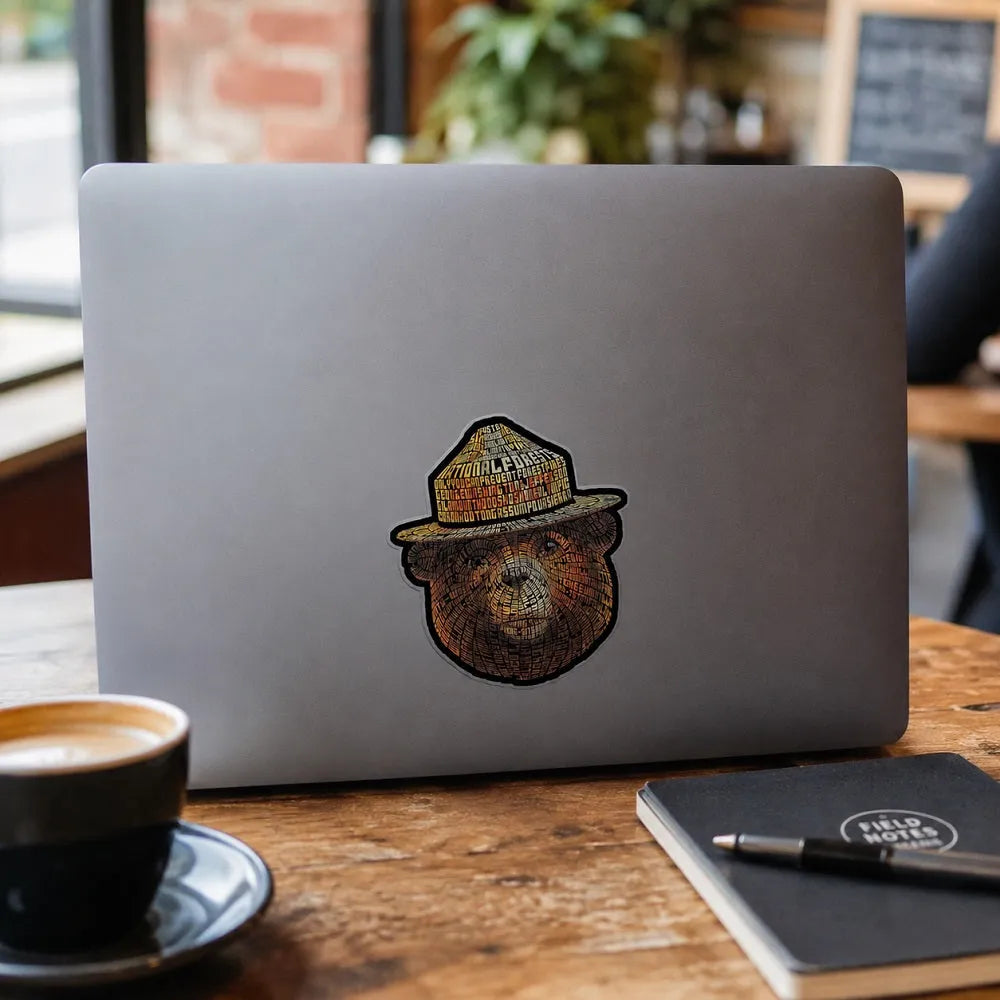 A silver laptop on a wooden table in a cafe setting. A coffee cup, a notebook with a pen, and a blurred cafe background are visible. The laptop has the Smokey Bear typography sticker on it.
