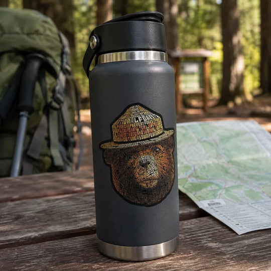 A dark gray insulated water bottle with a black lid sits on a wooden picnic table outdoors in a forest setting. The bottle features a detailed sticker of Smokey Bear.