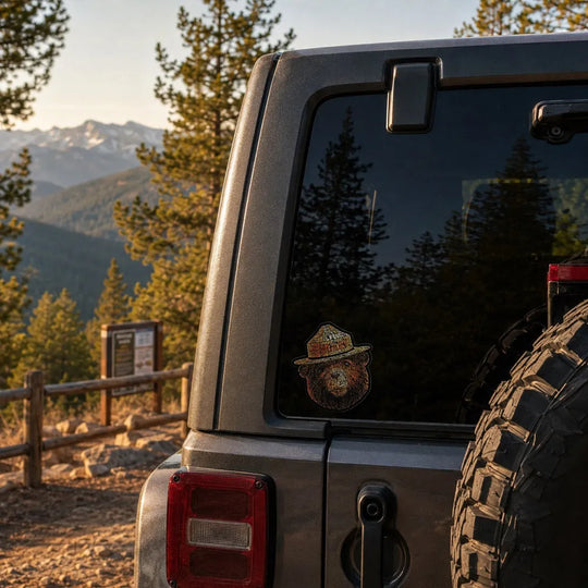 A close-up of the rear side of a gray four wheel vehicle parked outdoors on a mountain trail, Trees, mountains, and a wooden fence are visible in the background. The Smokey Bear typography sticker is on the corner of the car&