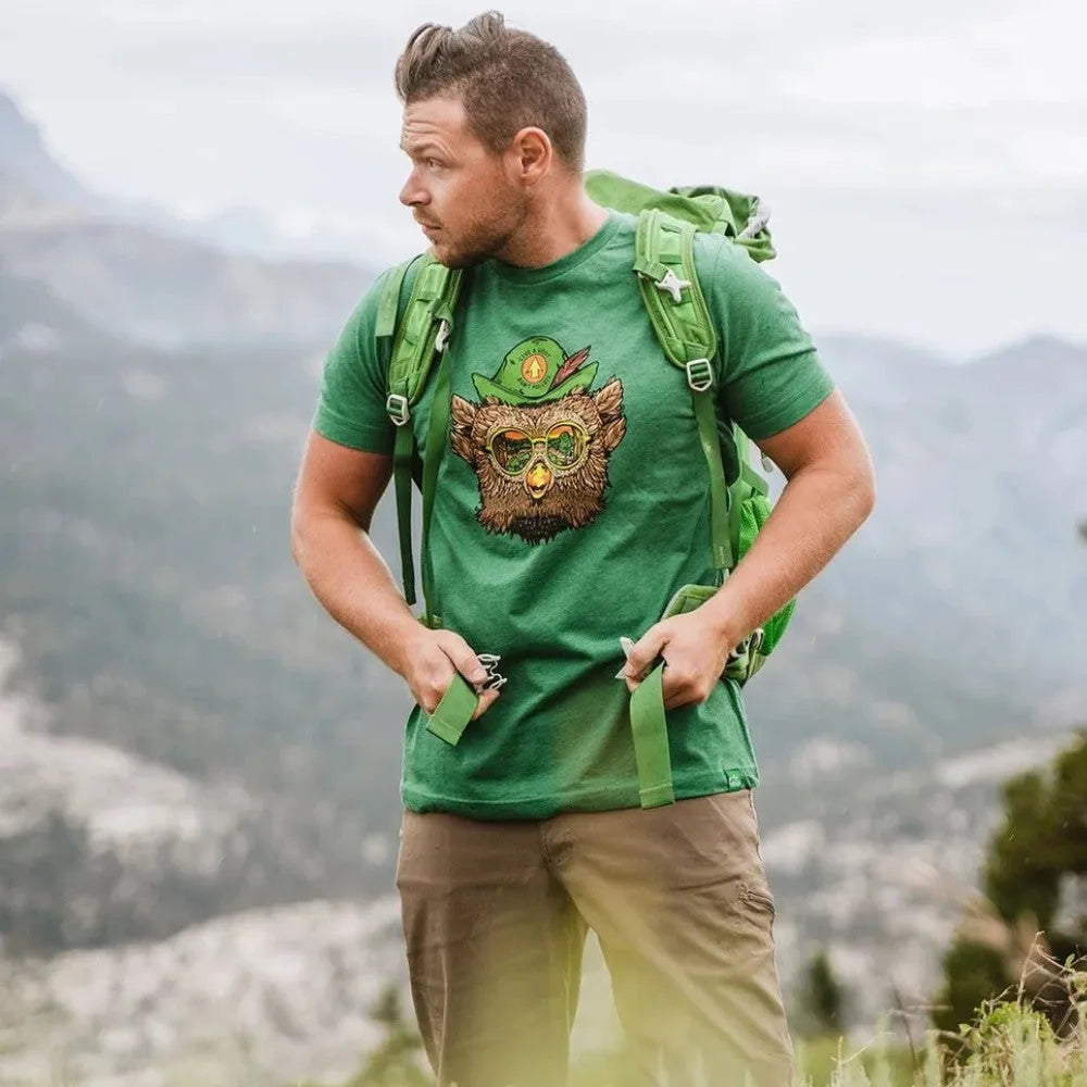 A man hiking outdoors wearing a green crew-neck t-shirt. The t-shirt has Woodsy Owl on it. The man is also wearing khaki pants and a green backpack and he is outdoors in the mountains.
