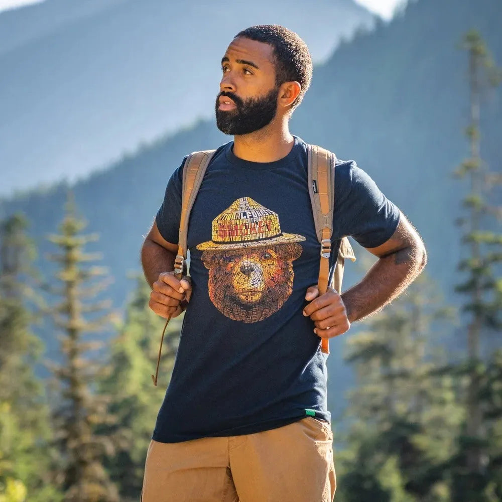 A man wearing a navy blue t-shirt with a colorful graphic of Smokey Bear on the front. The man is standing outdoors in a mountain forest setting with a backpack on.