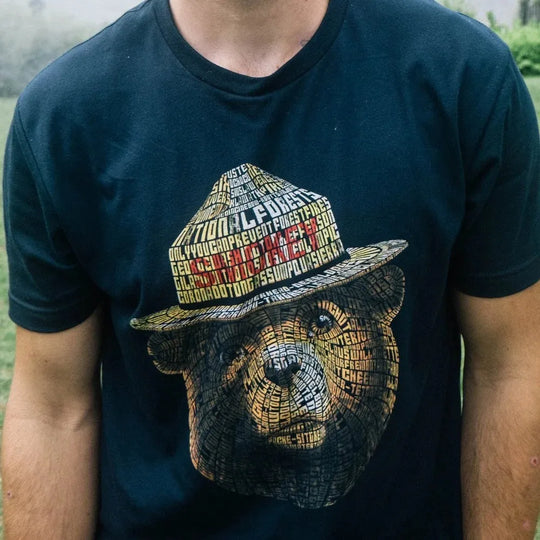 A person wearing a dark navy blue t-shirt with a large graphic of Smokey Bear wearing a hat. On a closer look, the image is made of up of different words, including the names of the national forests. 