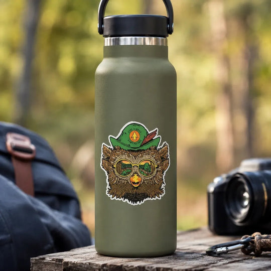 An olive green insulated water bottle with a black lid and handle with a sticker of Woodsy Owl wearing a pair of sunglasses. The water bottle is set on a wooden surface outdoors with a blurred forest background.