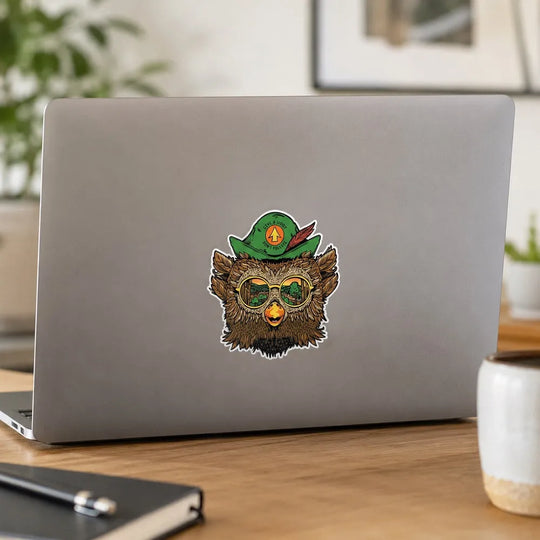 A die-cut sticker of Woodsy Owl wearing a green hat and round glasses, applied to the back of a silver laptop. The laptop sits on a wooden desk in a blurred home office setting with a notebook, plant, and mug visible.